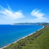 Una spiaggia lunga e un mare blu, con colline verdi in lontananza e cieli azzurri.