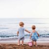 Due bambini guardano il mare sulla spiaggia, uno tiene un secchiello rosa.