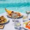 Un tavolo con piatti di pesce e formaggio, bicchieri di vino e d'acqua vicino a una piscina.