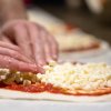 Mani che aggiungono cubetti di formaggio su una pizza con salsa di pomodoro.