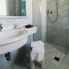 Bagno con lavabo bianco, doccia a vetro e asciugamani appesi.